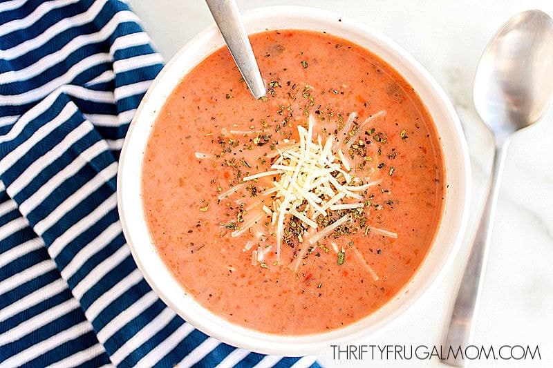 a bowl of homemade tomato basil soup topped with shredded parmesan cheese next to a spoon and dish cloth