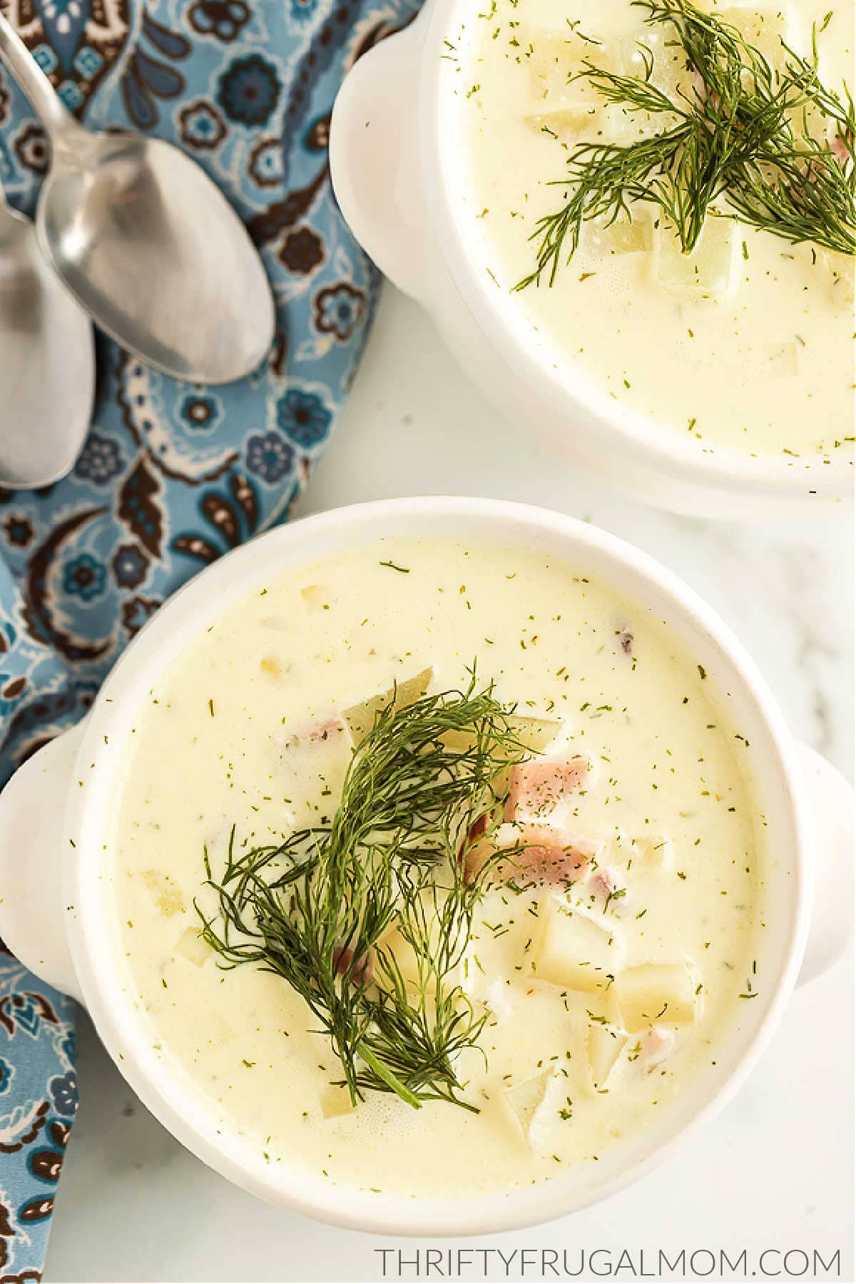 two bowls of cream cheese potato soup with ham topped with dill springs on a marble counter with blue cloth napkins