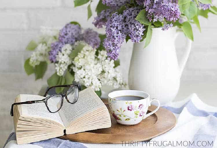 a book on a table along with a cup of coffee and vases of lilacs
