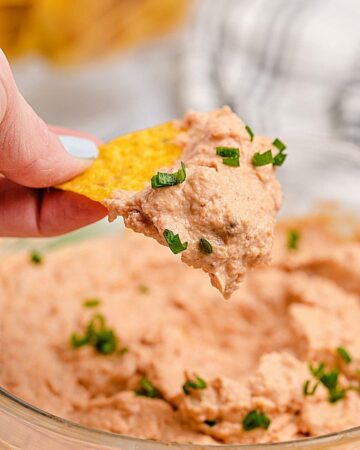 A closeup shot of a hand holding a triangle tortilla chip scooping easy refried bean dip out of a glass bowl.