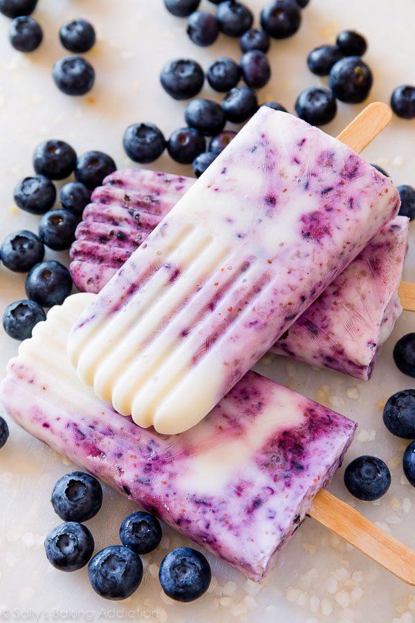cheap healthy snack of three blueberry yogurt popsicles lying on a counter with some blueberries scattered around