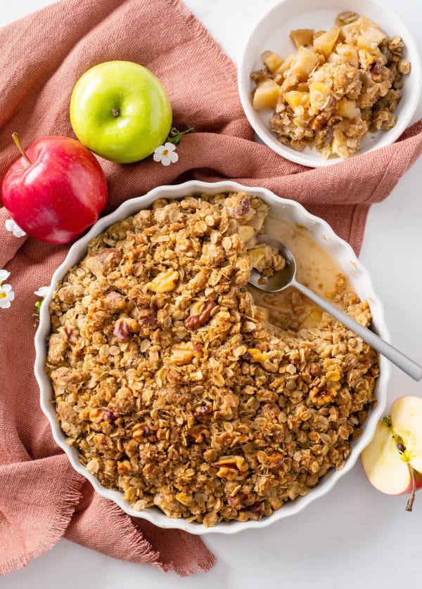 A round white baking dish full of no butter apple crumble with a spoon resting inside and fresh apples lying beside the pan.