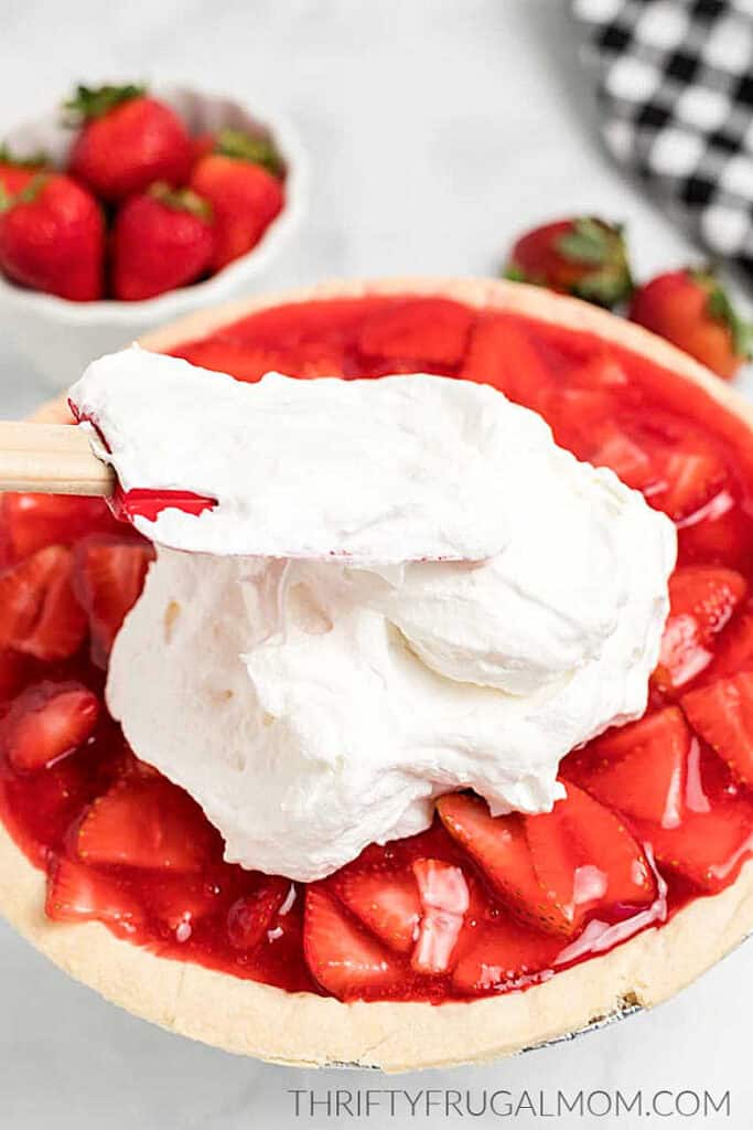homemade strawberry pie with jello being topped with whipped cream by a spatula