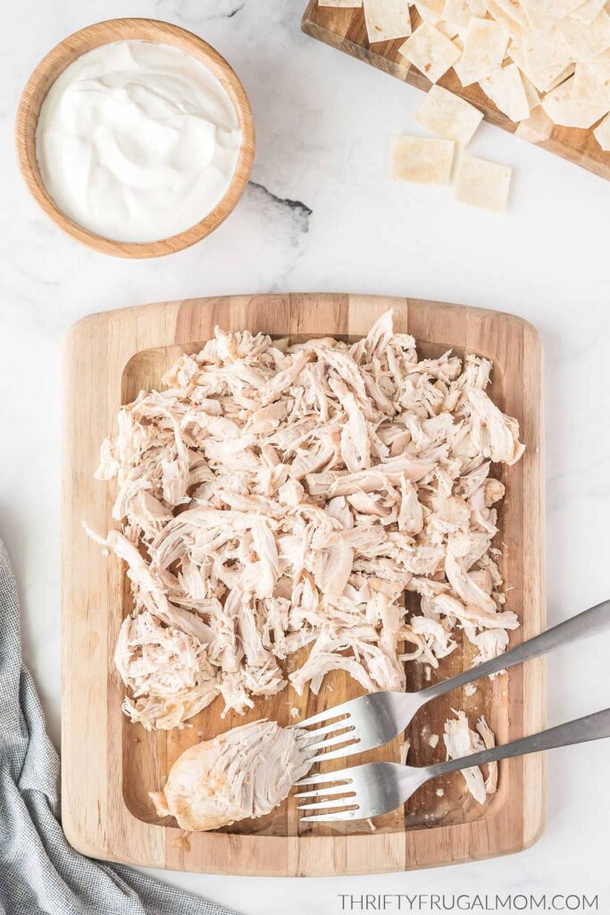 shredded cooked chicken on a cutting board with two forks next to a bowl of sour cream and tortillas that have been cut