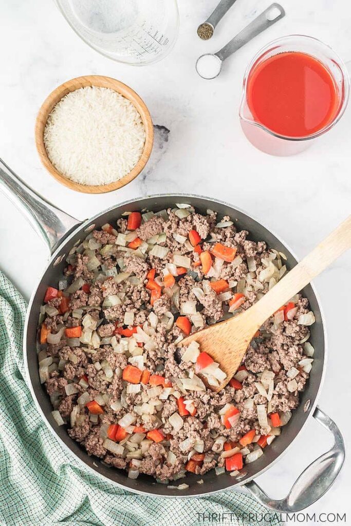 ground beef, onions, and peppers browned in a large skillet with wooden spoon next to green dish towel, tomato juice, and uncooked rice