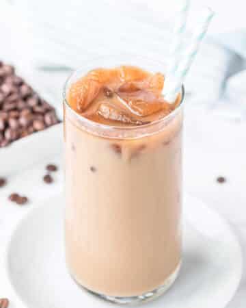 a vanilla iced latte in a glass with 2 blue and white paper straws next to coffee beans