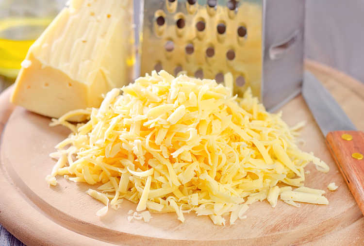 a pile of shredded cheese on a cutting board along with a knife, cheese grater and block of cheese