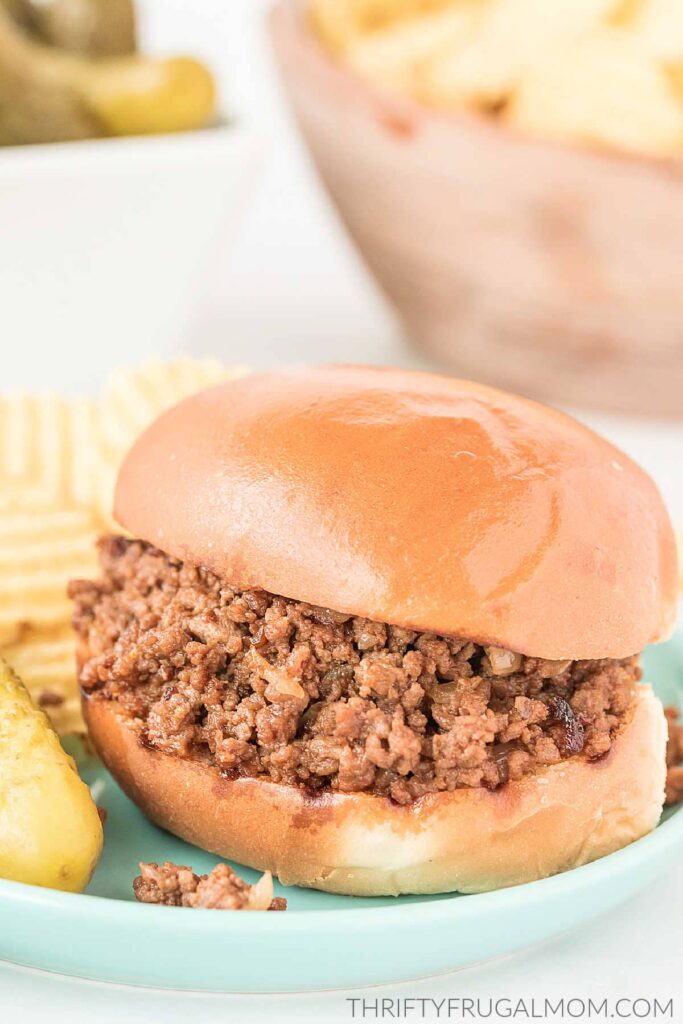 A sloppy joe sandwich on an aqua plate with chips and pickle.