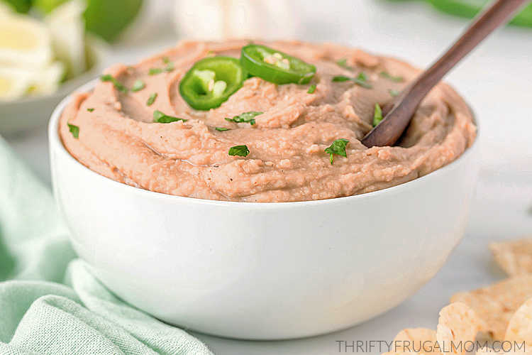 side view of homemade refried beans in a white bowl with a wooden spoon, topped with jalapenos.