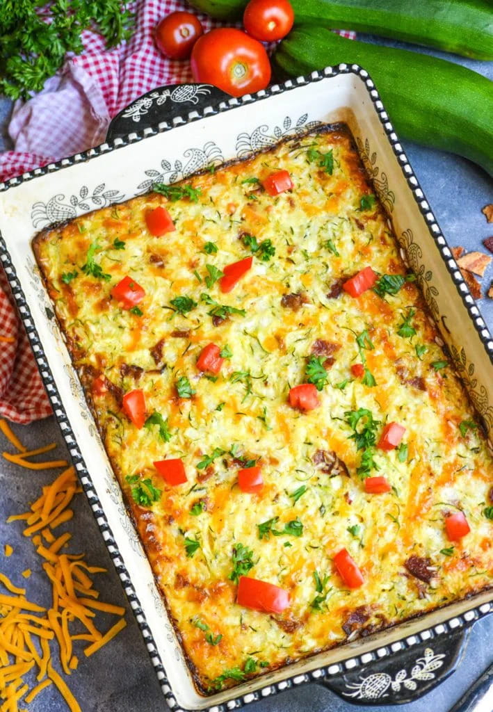 A large baking dish filled with Zucchini Bacon and Rice Casserole  on a blue countertop with shredded cheese scattered beside it.