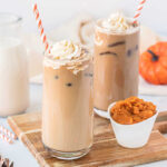 Two iced pumpkin lattes next to each other on a cutting board beside a bowl of pumpkin puree.