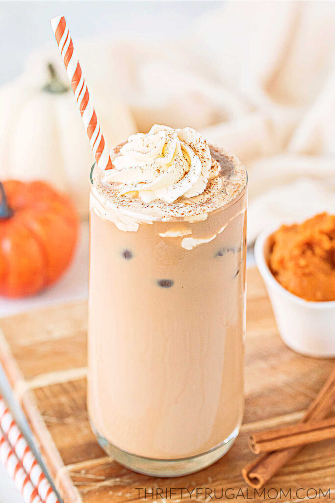 A homemade iced pumpkin spice latte in a glass topped with whipped cream with an orange and white paper straw.