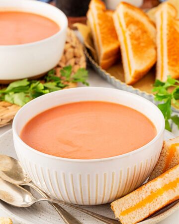 A white bowl full of easy Creamy Tomato Soup with grilled cheese sandwiches arranged around it and showing in the background.