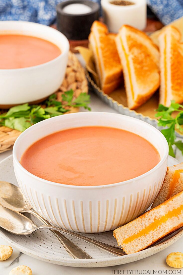 A white bowl full of easy Creamy Tomato Soup with grilled cheese sandwiches arranged around it and showing in the background.